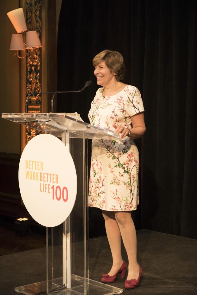 Founder and president Shifra Bronznick in a floral dress, smiling at a clear podium with a sign reading Better Work Better Life 100
