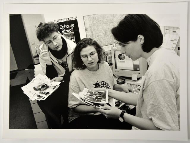 Three women in an office setting, looking at printed photographs. All three wear t-shirts with a pomegranate logo.