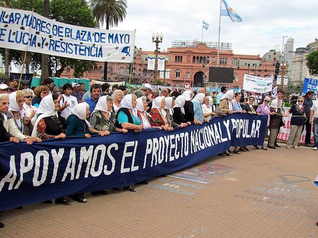 A crowd standing behind a line of women wearing white scarves and carrying a blue banner reading "APOY AMOS EL PROYECTONACIONALY POPULAR"
