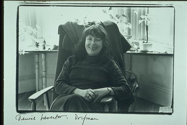 Denise Levertov sitting in a wooden chair, with potted flowers behind her