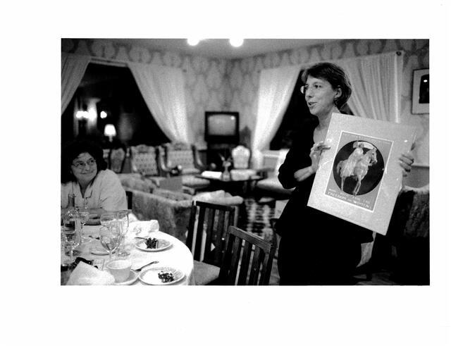 Gail Reimer standing beside a dinner table, holding up a print