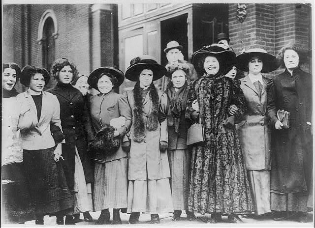 Nine women in long dresses, coats, and hats, with their arms linked, most smiling and laughing