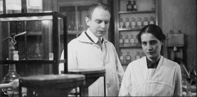 Lise Meitner and Otto Hahn, both wearing white lab coats, standing in a lab beside a shelf
