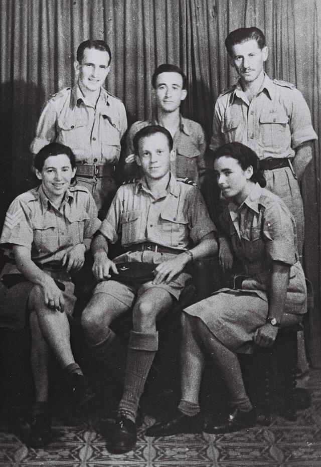 Six parachutists--four men and two women--pose for a portrait. Three stand behind the other three, seated.
