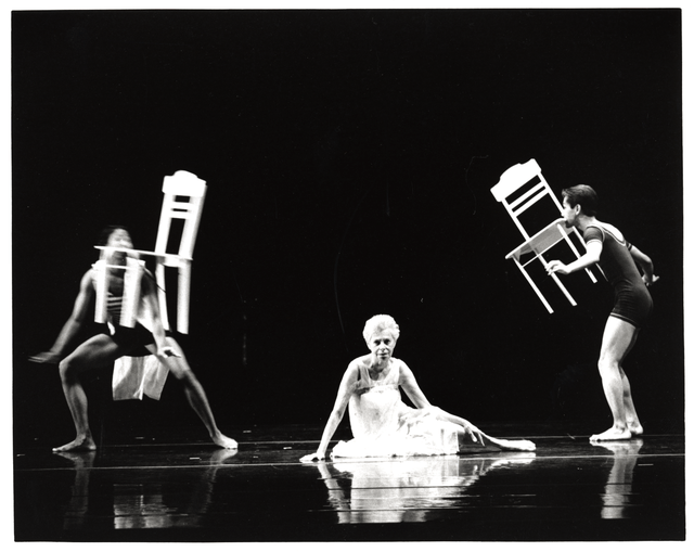 An older female dancer sitting on a stage, wearing a white dress, with two men in one-piece bathing costumes standing on either side of her, holding chairs with their teeth