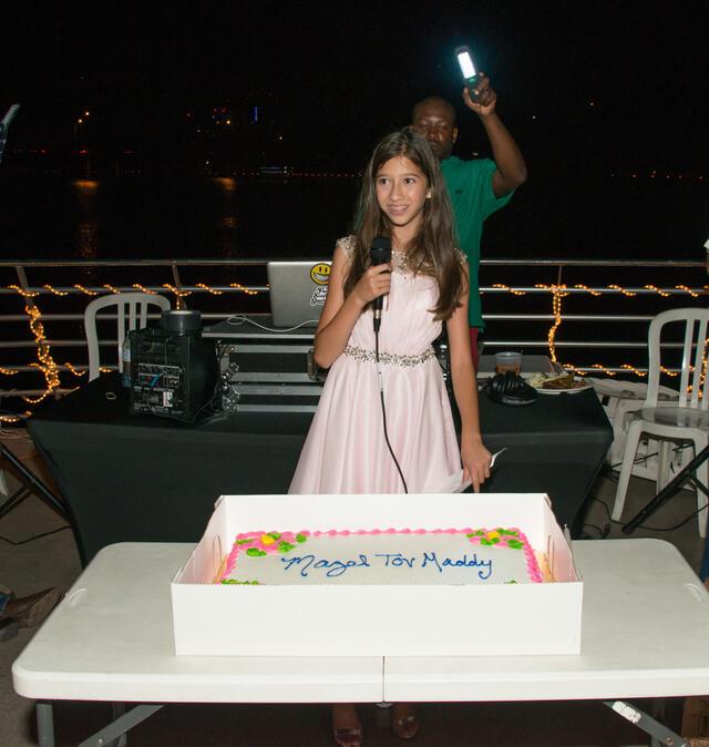 Maddy Pollack speaking at her Bat Mitzvah party, microphone in hand. Cake on a table in front of her with text "Mazel Tov, Maddy."