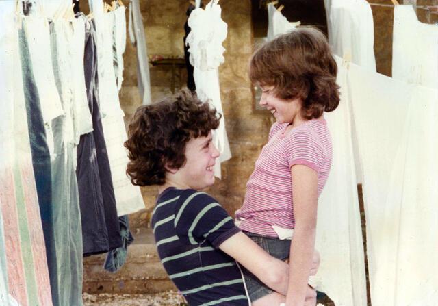 A boy with curly hair in a blue striped shirt lifting up a girl with shoulder length hair in a pink shirt. They are between clothes hang drying on lines.