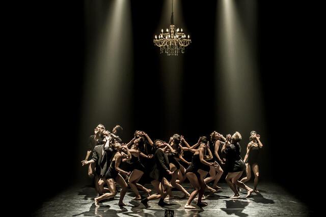 A large group of dancers in black leotards, and some in black jackets, dancing on a bare stage beneath a chandelier
