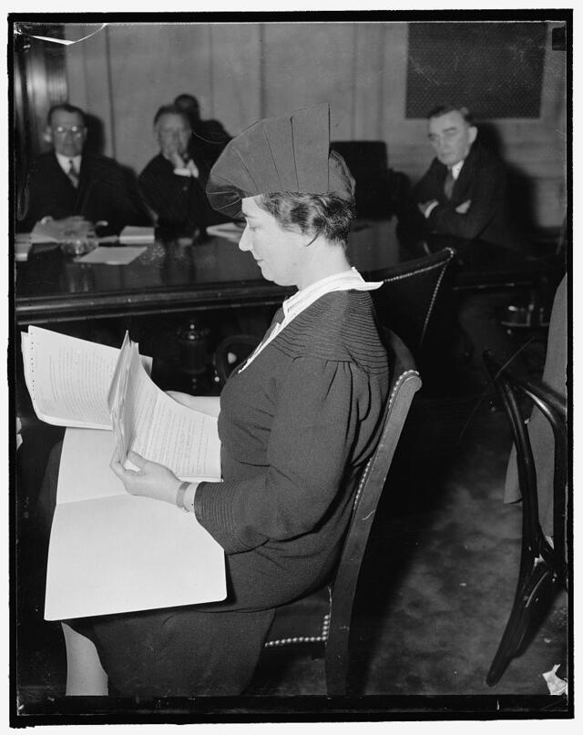 Dorothy Straus sitting a table, leafing through pieces of paper, with several men sitting in the background watching her