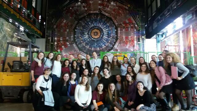 A large group of high school girls and some adults posing in front of the Large Hadron Collider