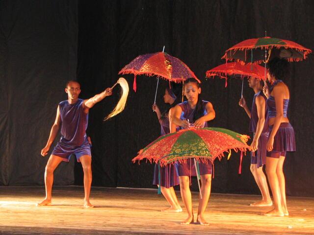A group of dancers on stage, all wearing loose blue/purple velvet shorts and shirts. Four are holding colorful red, red/purple, and red/green umbrellas, one holds a horse hair whip