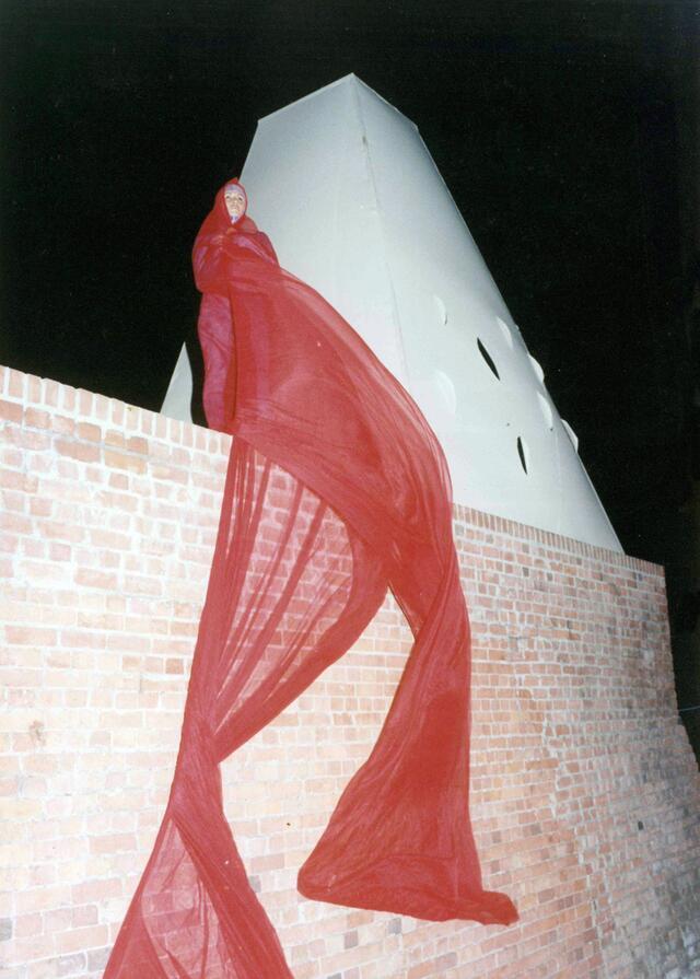 A dancer wrapped in a long, gauzy piece of red fabric standing on a brick wall beside a white tower