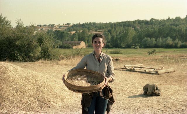 Ruth Behar standing in an open field with trees and houses in the background, holding a large wooden sieve