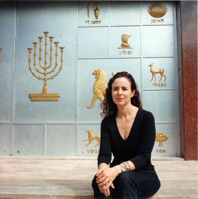 Ruth Behar sitting in front of a pale blue wall with embossed images of a menorah and symbols of the twelve tribes of Israel