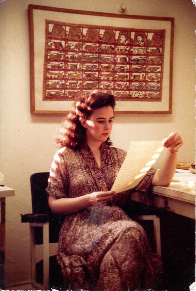 Ruth Behar sitting in a chair beside a desk, reading a piece of paper, beneath a large framed tapestry hanging on the wall