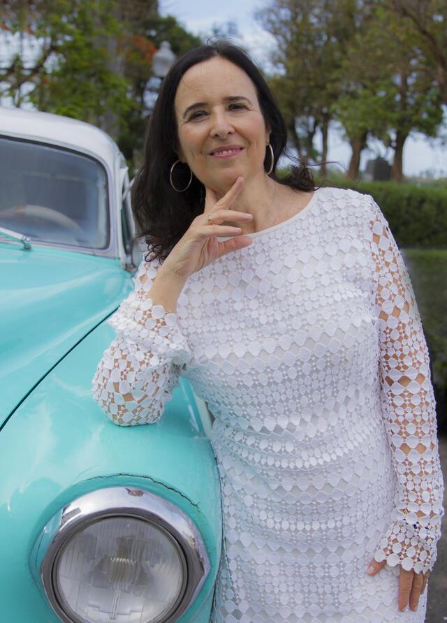 Ruth Behar, wearing a white dress, leaning against a turquoise car