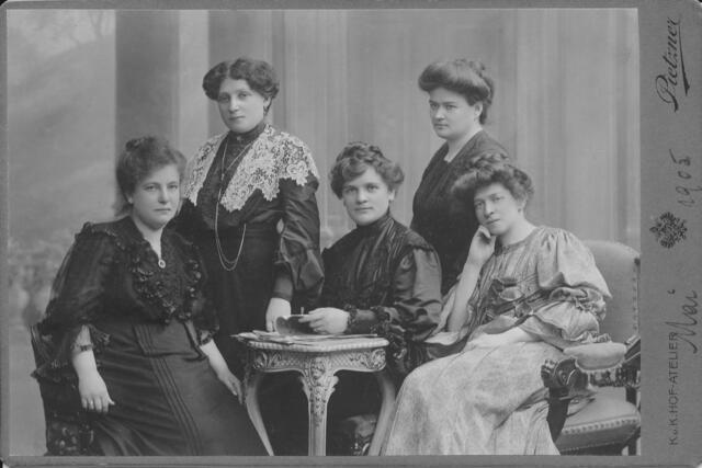 A group of five women in formal dresses, sitting and standing around a small end table