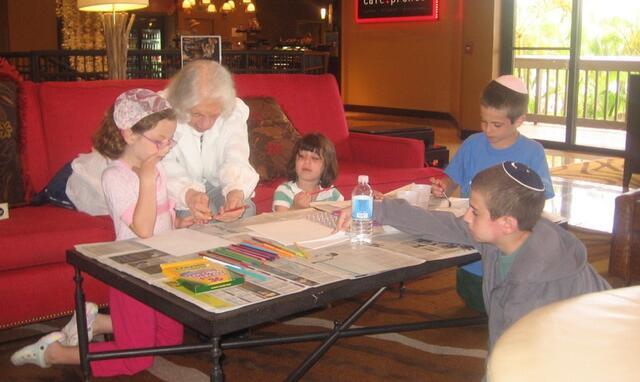 Grandmother coloring on paper with her grandchildren. They sit on and around a red couch.