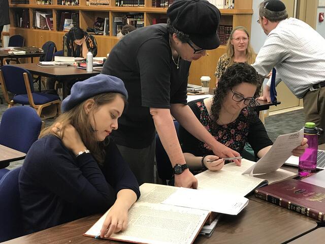 Several small groups of women studying at small tables, with teachers leaning over to assist them