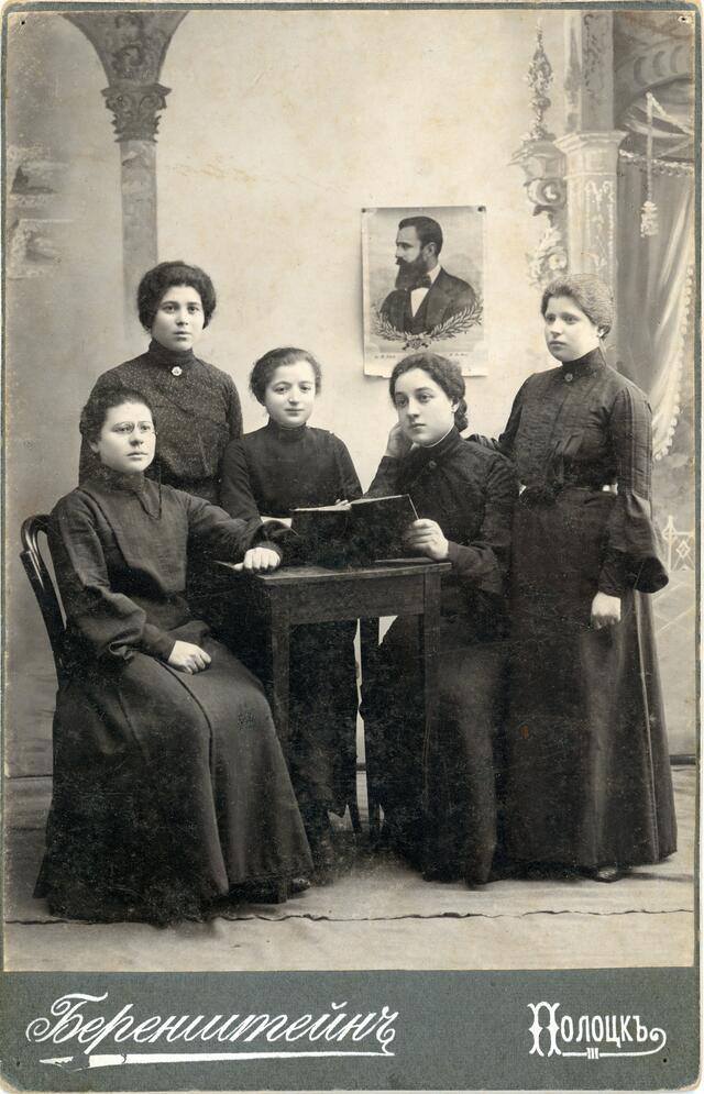 A group of women sitting around a small table with an open book, and a portrait of Theodor Herzl on the wall