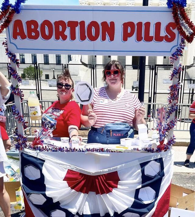 Two women stand at a booth with a sign that says "Abortion Pills"