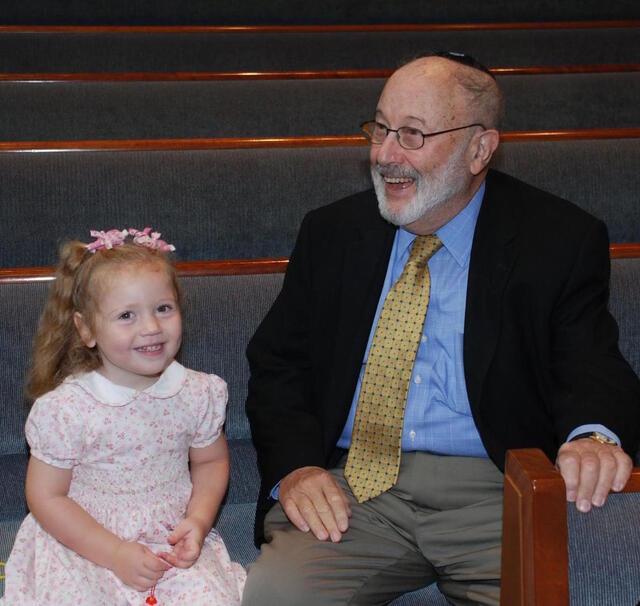 Six year old in pink dress seated next to elderly man dressed in a suit, sitting in pews at a synagogue
