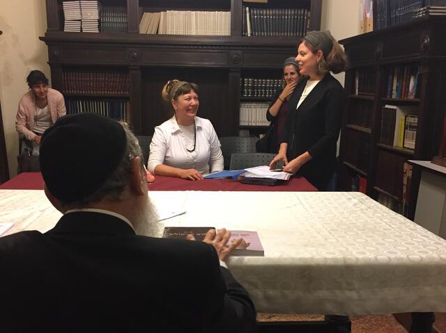 Woman seated at a table smiling at woman standing up, rabbi seen from the back. 