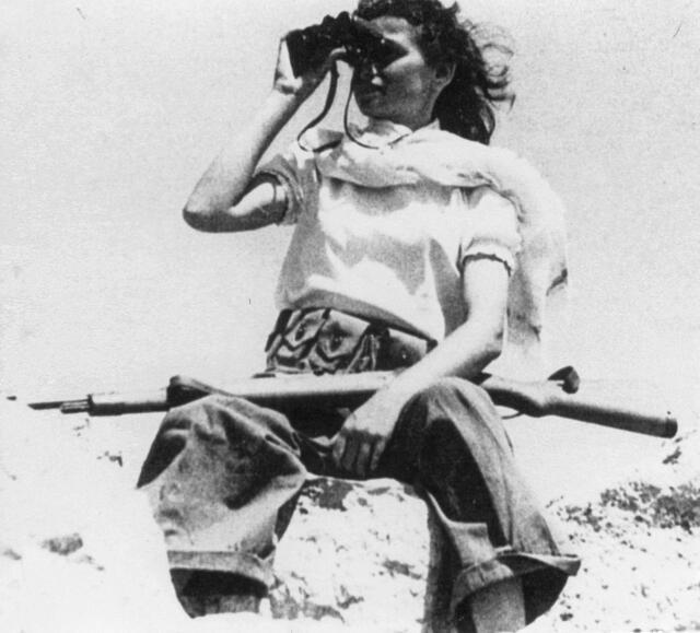 A young woman sits with a rifle across her lap and looking through binoculars.