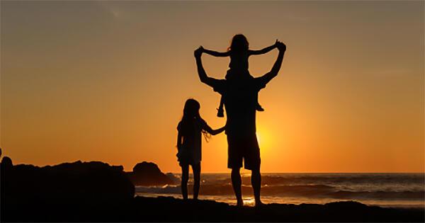 Silhouette of children with adult in front of beach sunset, one of the children is on the adult's shoulders and the other is next to them.