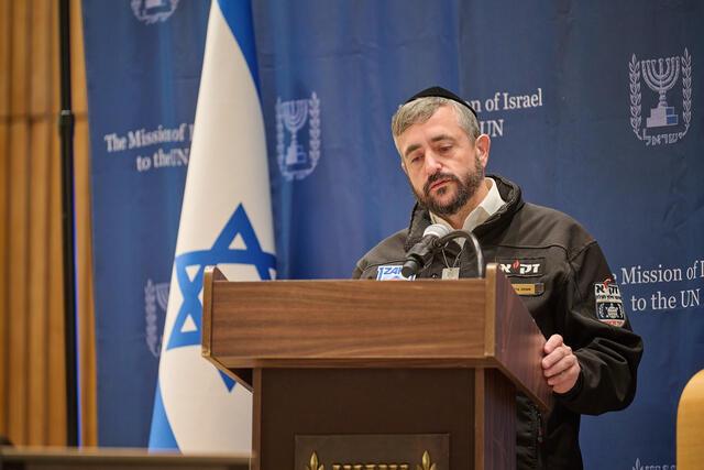 Man with kppah and beard speaks at podicum with Israeli flag in background