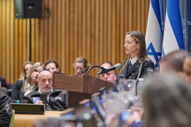 Woman in police uniform speaks at a podium, Israeli flag in background